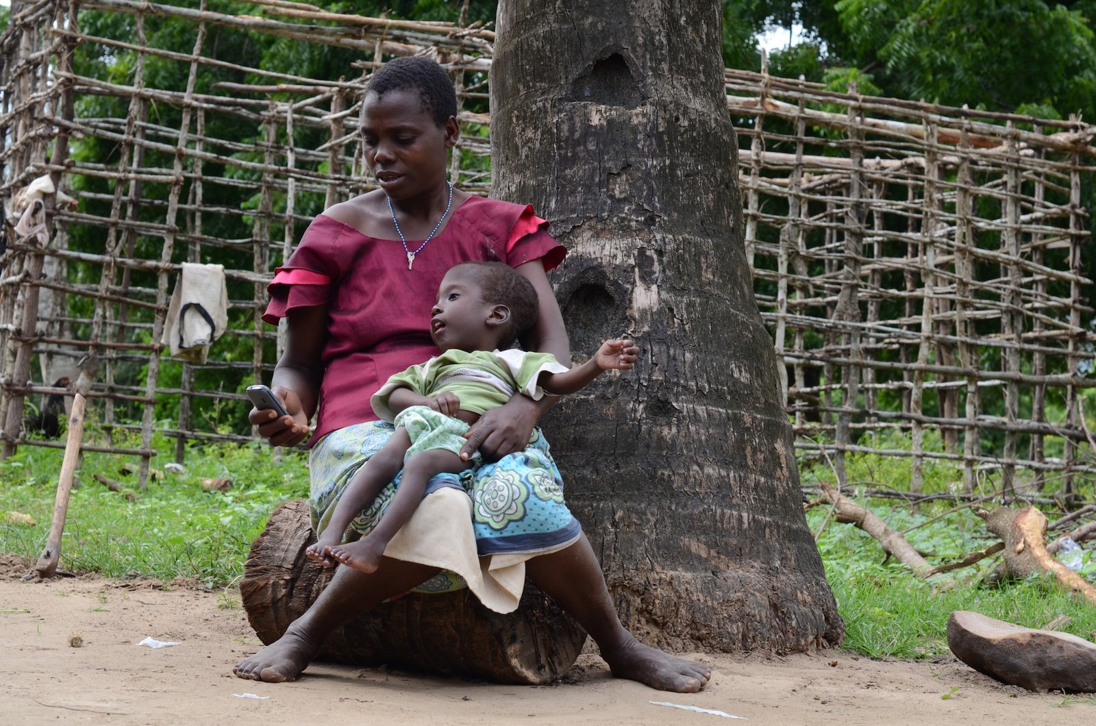 A mother sits on a log against a palm tree with her profoundly disabled son in her lap. She is looking at her mobile phone.